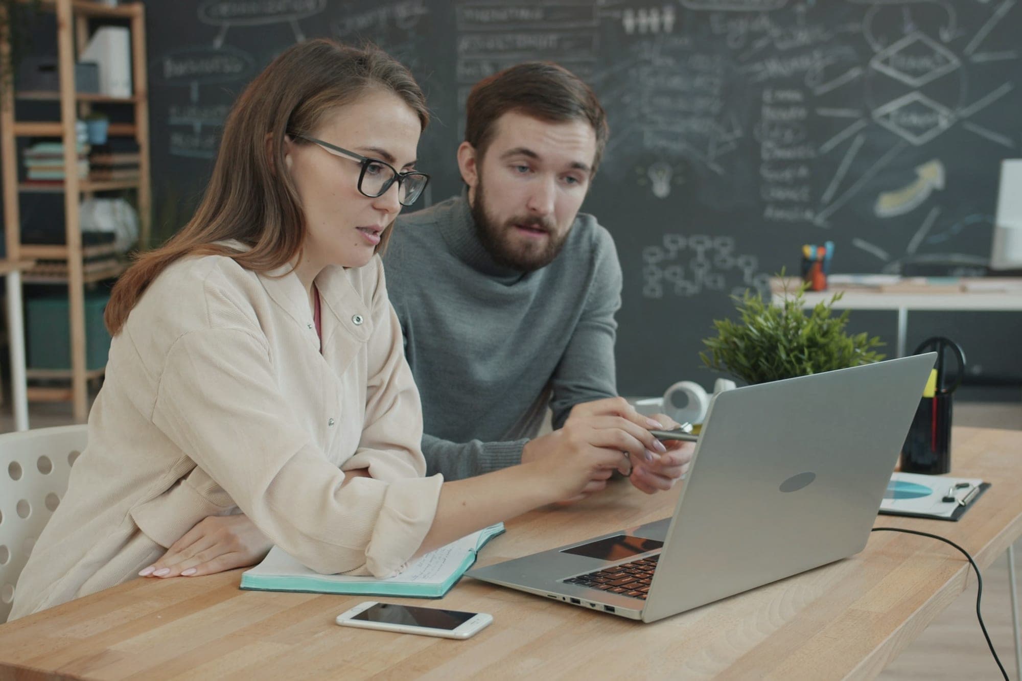 Two colleagues at a laptop, one pointing at the screen while the other follows along