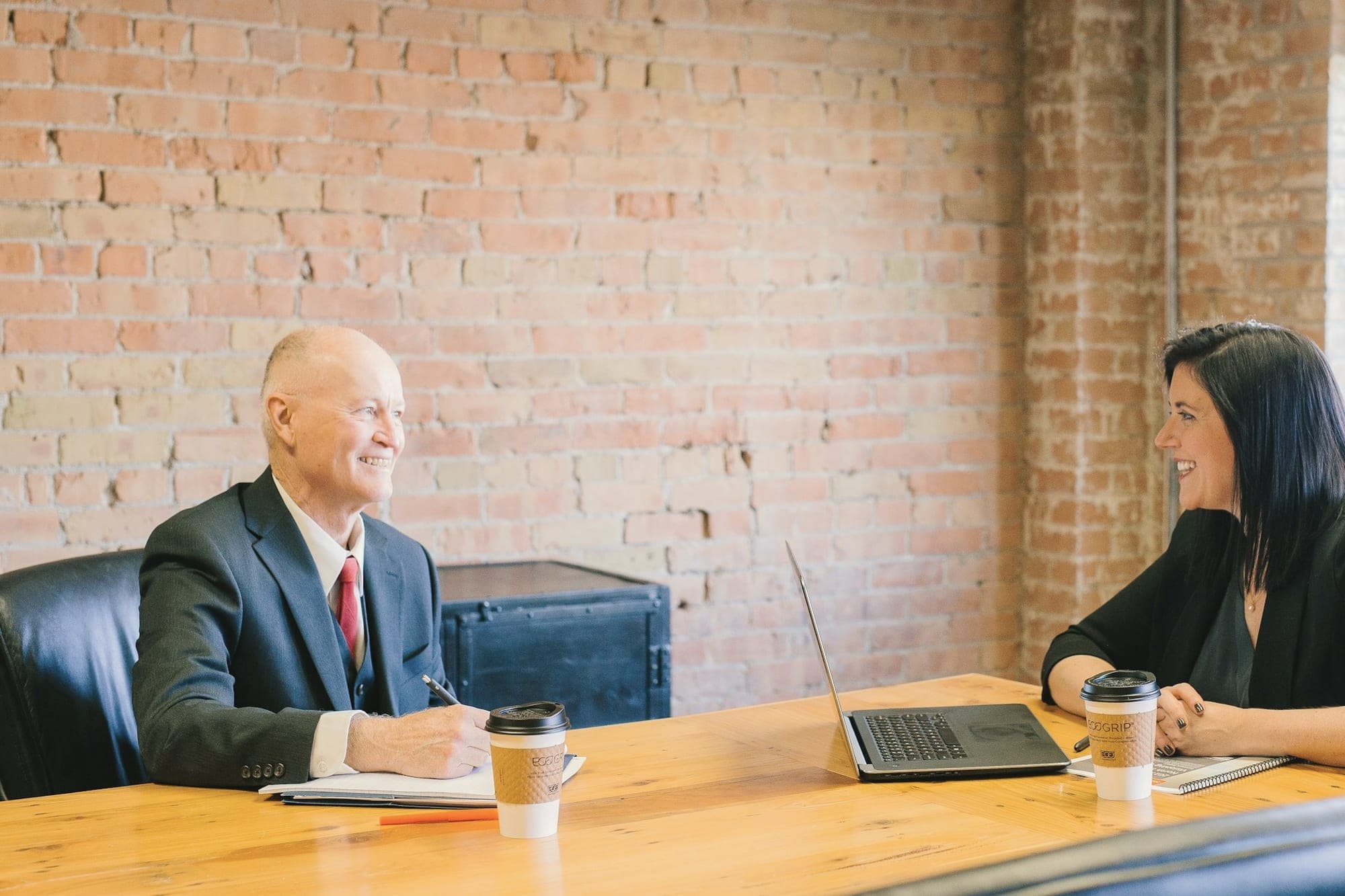 A man and a woman talking across a wooden table with coffee cups and a laptop