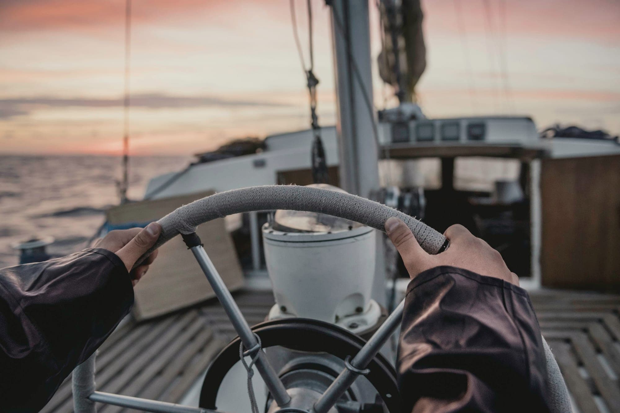 Hands on a boat's steering wheel, the sea and a pink sky ahead