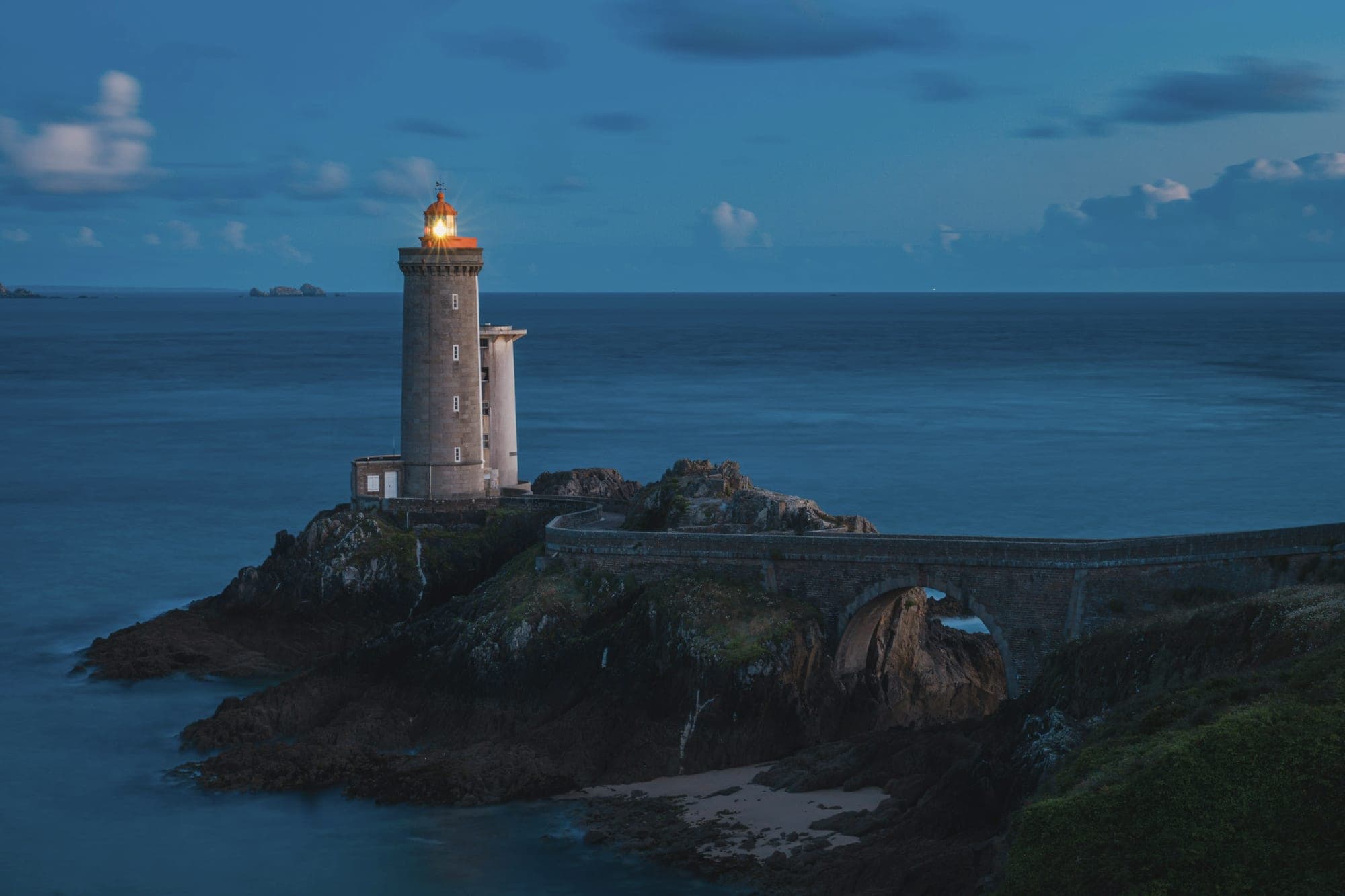 A lighthouse lit at dusk on a rocky point above the sea