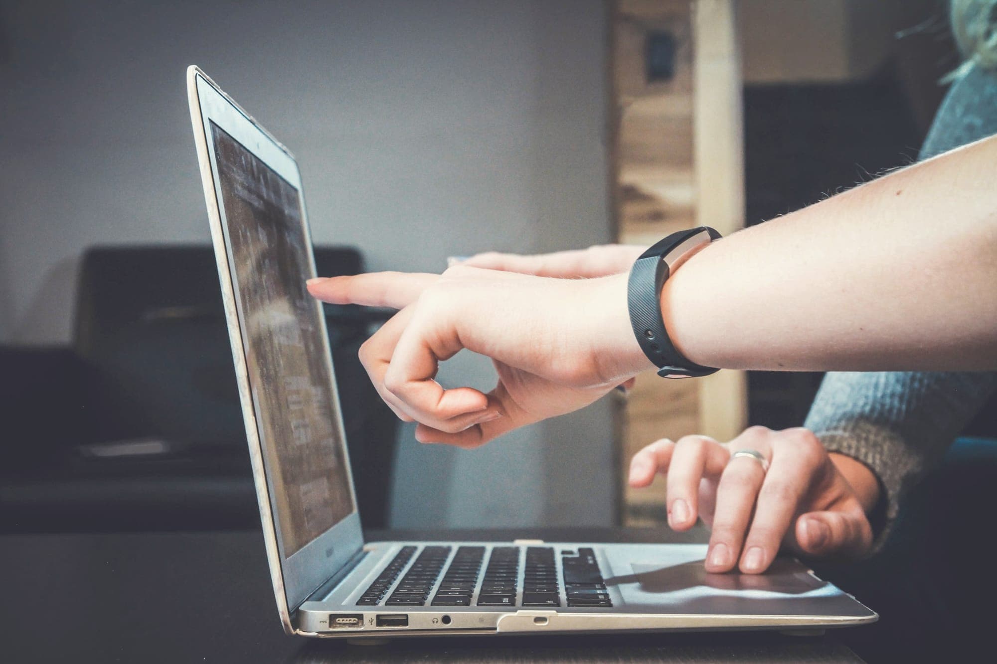 A hand pointing at a laptop screen while another hand rests on the trackpad